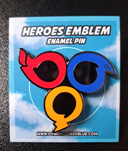 A photograph of a small enamel pin featuring a stylized design inspired by the team emblem from Sonic Heroes, attached to a backing card with an open blue sky, as a nod to the game's boxart.