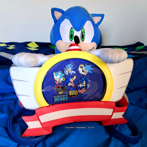 A photograph of an ita bag shaped like the winged emblems from various Sonic games, on a bed with a blue comforter and a Sonic-themed throw blanket behind it. A large Sonic plush is sitting behind the bag with his hands on the wings of the bag. The window of the ita bag has various Sonic pins inside it.