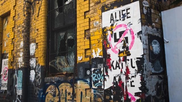 The side of a run down brick building, painted yellow on top, and black below - covered in graffiti and anti-fascist/anti-nazi posters that read “Alife Katsu.” The posters appear worn and torn and fit the decaying nature of the rest of the building. To the left of center, a window pane with broken glass sits behind a metal barrier