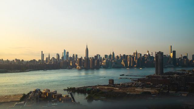 The New York City skyline at dusk. The left side of the image still showing a bright orange light hitting the distant NYC buildings, a river and docks in the foreground of the image