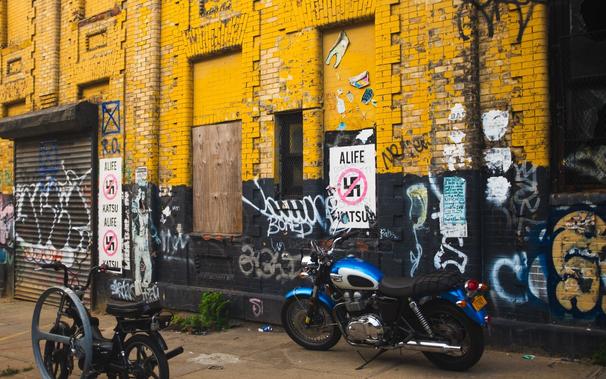 A blue and silver motorcycle sits in front of a yellow and black brick building. Above the moto is an anti-fascist / anti-Nazi poster with a cross buster over a swastika that reads “Alife Katsu”. Several posters can be seen further down the wall. The building os covered in graffiti