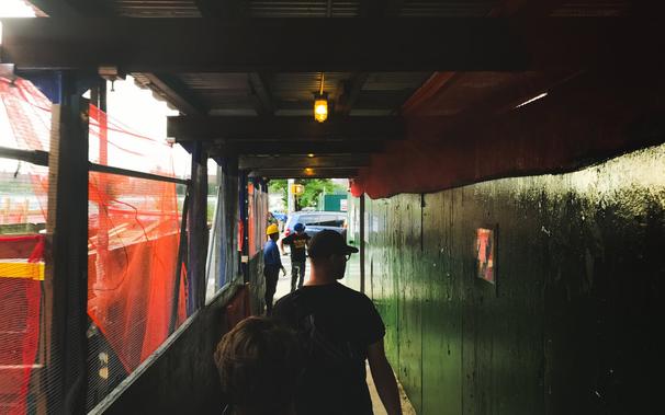 People walk ahead on a covered sidewalk - a green wall on their right, orange mesh covering from a construction zone on the left. A person in center frame is framed within the light coming from outside the walkway. Orange incandescent lights shine above the walkers