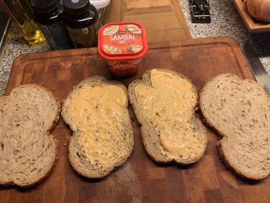 Slices of whole grain bread on a wooden cutting board. Two of the slices have a spread on them, which appears to be a creamy, orange-colored sambal cheese spread. The container of the spread is placed above the slices, labeled "Jumbo Sambal 48% Smeerkaas." In the background, there are bottles of cooking oil and other condiments, as well as a wooden box with onions