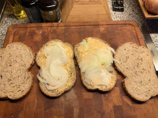 Slices of whole grain bread are laid out on a wooden cutting board. Two of the slices have a layer of orange cheese spread and thinly sliced onions on top. The other two slices are plain. In the background, there are bottles of cooking oil and a container of peppermint softgels. A knife is placed on the right side of the cutting board