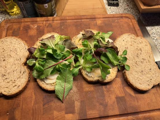 Two slices of whole grain bread are topped with fresh mixed greens and thinly sliced onions, ready to be made into sandwiches. The bread slices are placed on a wooden cutting board. In the background, there are some kitchen items like a bottle of oil, a can, and a container of peppermint pastilles. A knife is partially visible on the right side of the cutting board