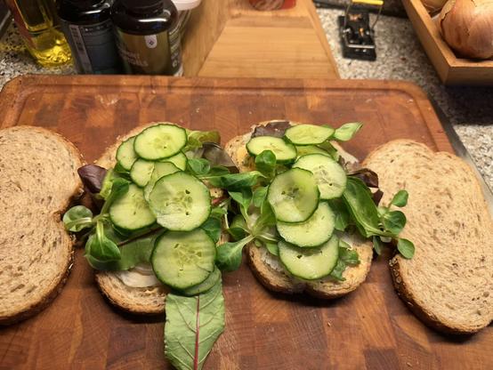 Two slices of whole grain bread on a wooden cutting board, each topped with fresh green lettuce leaves and several slices of cucumber. The sandwich is open-faced, with two additional slices of bread placed nearby, ready to be used as tops. In the background, there are various kitchen items, including bottles of oil and spices, a binder clip, and a wooden box containing onions