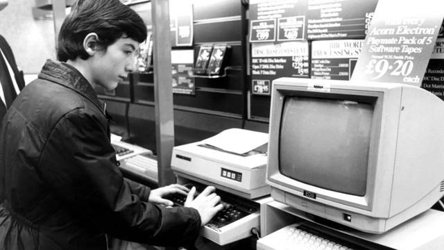 Black and white photo of a young man typing on a BBC Micro next to a CRT monitor and other 1980s computing equipment