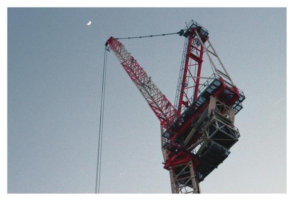 Color photograph of a red and white construction crane, taken at dusk. The perspective from below shows the counterweights and the luffing jib pointing towards the half moon, which can be seen in the left part, at the top of the landscape-format photo.