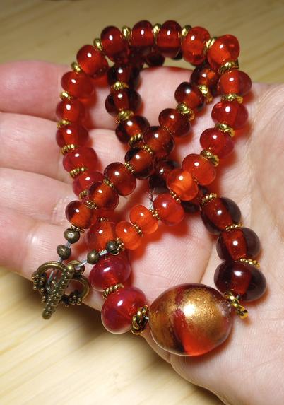 Image of my handmade lampwork hot glass necklace in transparent red and topaz, with brass spacers and toggle, and a large focal bead at the center, enhanced with gold colored mica fused to the glass, held in my open palm, set against a light wood background.