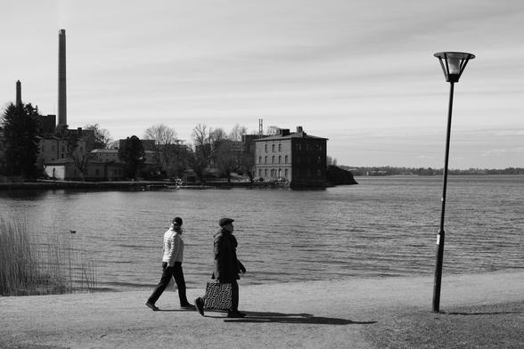 A black and white photo of two people walking along the coastline in Lapinlahti, Helsinki, on a sunny spring day. There is an old hospital in the background and and a street light on the right.