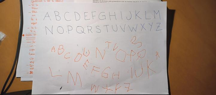 Folio horizontal en el que se ve en letras azules mayúsculas escrito el abecedario en la parte de arriba y en la parte de abajo dispersas sin orden (empezó en orden pero le faltó el espacio) en color naranja la copia de esa letras hecha por un niño de 3 años y 8 meses.