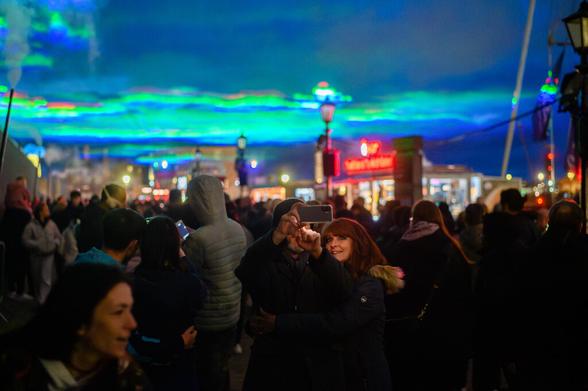 A crowd of people gathers at night to watch a light show at the Royal Albert Dock in Liverpool, UK. The light show projects colorful aurora-like patterns onto the sky. One couple takes a photo with their phone.