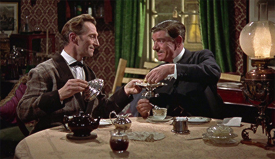 This is a photo of Peter Cushing as Sherlock Holmes and André Morell as John Watson. They're sitting across from one another at a table at teatime.