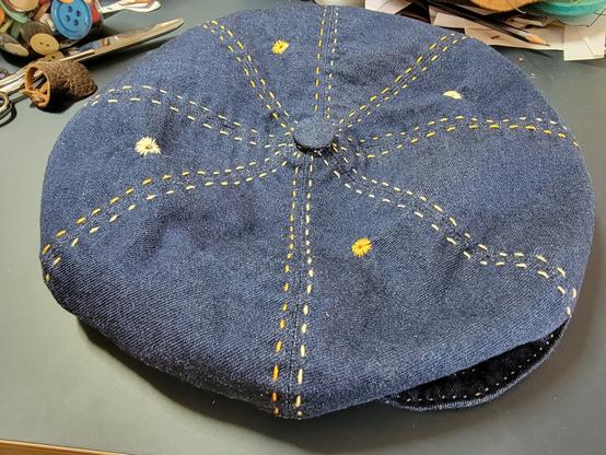 A dark blue denim newsboy cap with variegated yellow stitching and four yellow eyelet vents.