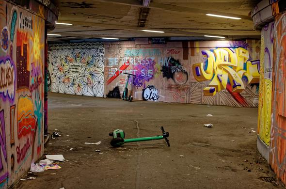 Eine unterirdische Rad- und Fußgängerpassage. Die Wände voller Graffiti. Ein türkisfarbener Scooter steht an die hintere Wand gelehnt, einer liegt im Vordergrund auf dem Boden. Deckenleuchten erhellen die Passsage spärlich-
