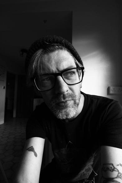 me, a bearded white man with tattoos wearing glasses, dark short-sleeved t-shirt, and a beanie