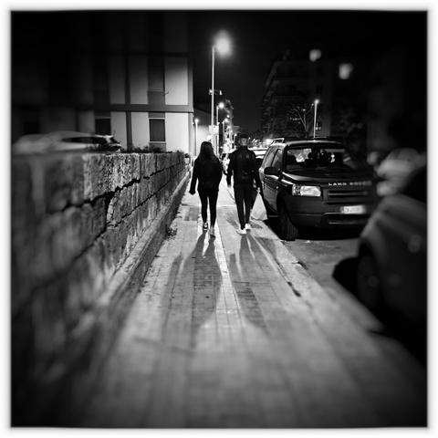 Una coppia passeggia in un marciapiede in città illuminato da un lampione, macchine parcheggiate a destra e muretto in pietra a sinistra