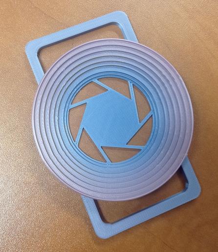 Photo d'un porte capuchon d'objectif imprimé en 3d de même couleur que le capuchon. L'objet est un genre de disque avec un dégradé de sa hauteur vers son centre, qui permet d'accrocher des capuchons de tailles différentes.