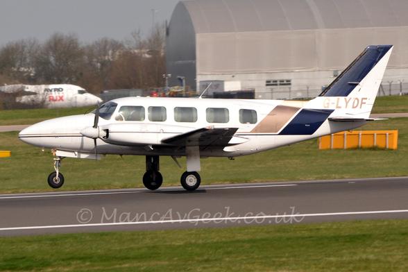 Side view of a white, twin propellor-engined BizProp rotating as it takes off, moving from right to left with it's nose-wheel lifted off the ground and main wheels just lifting.
There is a thin gold and blue stripe running along the body, turning up into the tail and getting wider, the gold stripe wrapping over the top of the body, the blue stripe forming the front of the tail.
Ther registration "G-LYDF" is on the lower part of the tail, reflecting low but strong sunlight.
The plane's pilot can be seen through the cockpit side window, but the dark cabin doesn't reveal any passengers.
Green grass fills most of the foreground, with more grass leading up to a large grey hangar on the right, and the forward fuselage of a white jet airliner with blue and red "Avro RJX" titles on the side on the left of the frame.
Leafless trees and grey sky fill the rest of the frame.