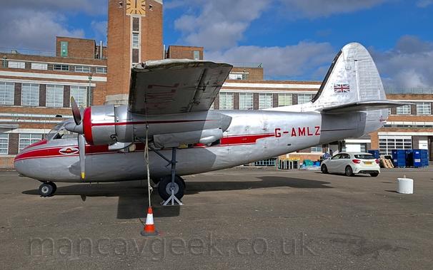 Side view of a silver, high-winged, twin propellor-engined light transport aircraft parked facing to the left.
There is a red stripe running along the body, with red registration "G-AMLZ" in the rear fuselage, the registration also being visible under the left wing.
There is a British flag on the tail.
There is an orange and white traffic cone on the ground under the wing, attached to the underside of the wing by a rope.
The main wheels are lifted off the ground, held by triangular metal frames.
In the background, a large, brown brick building with lots of windows stretches across the whole frame, with a clock tower behind the wings rising up into the blue sky.