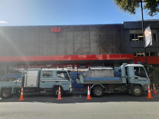 A new looking two-story building is close to being finished building. There are two service vehicles parked outside and many workers around the area. The building has the Westpac logo and red colour design on it.