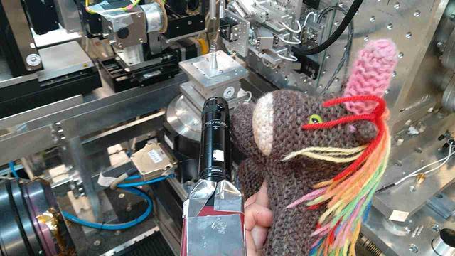 Max, das bunte Strickhorn, hilft dabei, eine der Kameras im 3D-Röntgenmikroskop auszurichten. Mittig vorne ist die Kamera mit einer Bleischürze als Strahlenschutz; sie ist auf einen Probenturm ausgerichtet. Auch im Bild sind Motoren, Mikroskope, Röntgenoptik, -detektor, Gedöns.