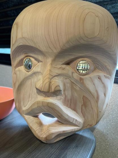 A close-up of a hand-carved wooden cedar mask, of Indigenous Coast Salish artistry. The mask has a smooth finish with visible wood grain. Its eyes are inlaid with iridescent abalone shell, and it features a wide, open mouth. The mask rests on a dark grayish-blue table.
