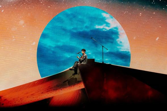 Wide shot of Seungmin sitting on the edge of a stage set object, playing a guitar. Behind him is a giant red circle reminiscent of a moon, surrounded by a red-tinged sky