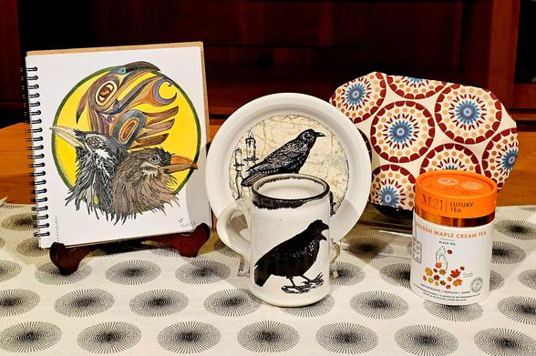 Tea tableau with a raven theme. There is a painted pottery mug with a raven design made by an artisan in Alaska, a Fiestaware plate with a raven image, and hand colored artwork with depictions of ravens including one in the Tlingit style. The tablecloth and tea cozy have spiral designs. The tea is Golden Maple Cream black tea.