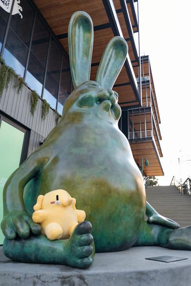 Photo of the Creature Mastodon stuffed toy sitting on a giant, unusual rabbit sculpture outdoors in Los Angeles during a cloudy afternoon. The Creature's sitting on the lap of the “Be Hare Now” sculpture by the Haas Brothers, which is sitting on a concrete platform, as they both stare up towards image top-right.