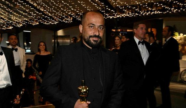 Hamdan Ballal in a black suit with his Oscar at 2025's Academy Awards. Some kind of Oscar reception is going on behind him,