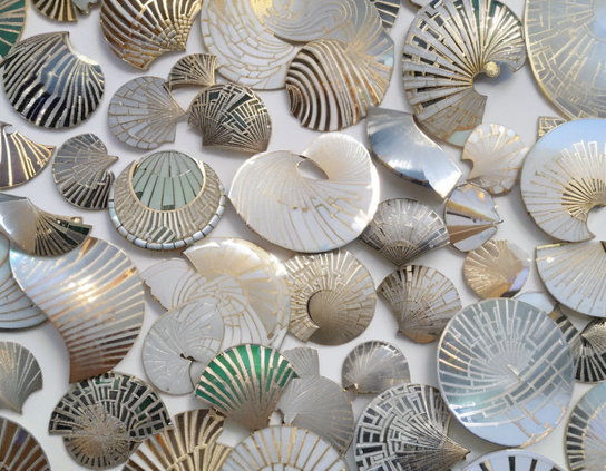 Assorted stylized metallic mollusk shells in gold, silver, white, and some green, with geometric inlay patterns, on a light background.