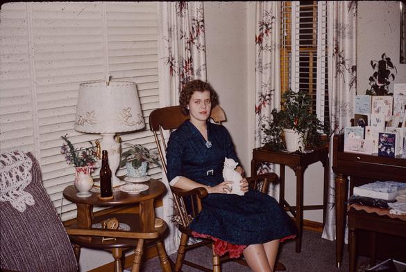 Young woman, probably late teens to mid twenties, sits in a padded rocking chair. She's in a royal blue dress with a broach and a black belt. She's holding a white ceramic cat on her lap.