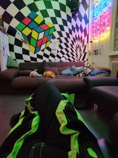 Photo of a couchcorner in a hackspace. 
The wall behind the couch has a large rubiks cube painting on it with some optical illusion. 
On the wall on the right of the couch are a bunch of RGB LEDs lit up in the colors of the Pansexual pride flag (pink, blue and yellow).
In the foreground my legs in green ravepants can be seen.