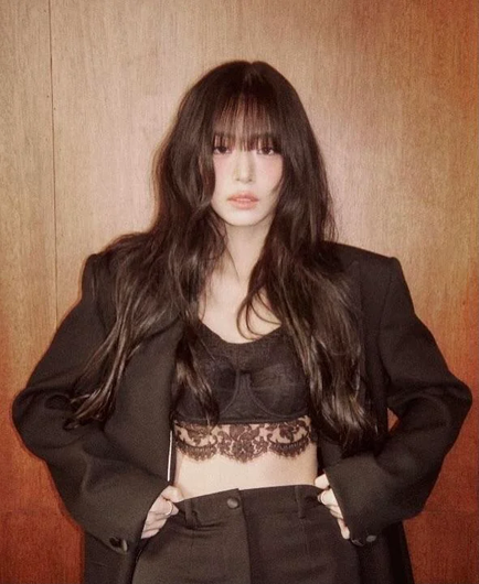 The image features Kpop idol  Chaeyoung of the group Fromis_9 standing against a wooden background. She has long, dark hair with bangs and is wearing a black oversized blazer over a black lace lingerie style crop top, which reveals a portion of the midriff. The person is also wearing black pants. The blazer has a relaxed fit, with the sleeves slightly oversized, and her hands are placed on the hips, with fingers tucked into the waistband of the pants. The lace on the crop top is detailed and adds a delicate contrast to the otherwise solid black outfit. The lighting is soft, highlighting the textures of the clothing and hair.

Provided by @altbot, edited by @jemmesedi