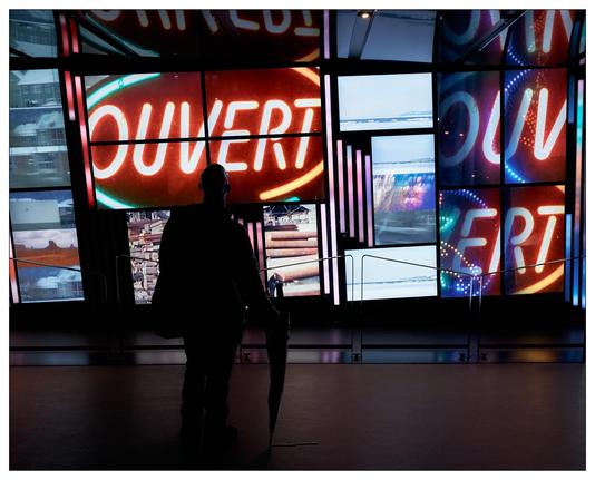 A silhouetted figure of a person in the foreground, standing indoors and possibly watching or moving towards a large display of screens. These screens show the word “OUVERT” prominently in a neon style, which means "open" in French. The display has an assorted set of visual elements, including what appears to be urban scenes and abstract patterns, creating a vibrant and dynamic atmosphere. The lighting and composition suggest a modern and possibly artistic venue.