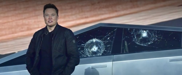 A photograph of Elon Musk with that self-satisfied rich-kid Nazi sneer that he often sports, standing beside a Cybertruck with multiple broken windows.