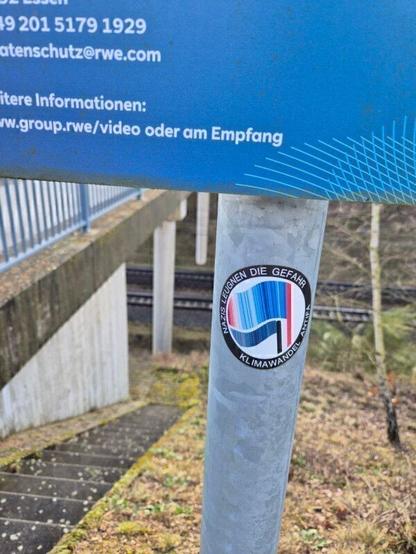 Das Bild zeigt einen Teil einer Treppe, die zur Hambachbahn hinunterführt. An einem Metallpfosten neben der Treppe ist ein blaues Hinweisschild angebracht, das Kontaktdaten und Informationen zur dort stattfindenden Videoüberwachung enthält. Auf dem Pfosten ist ein runder Aufkleber befestigt. Der Aufkleber zeigt ein stilisiertes Flaggenmotiv mit warming stripes und die umlaufende Aufschrift „Nazis leugnen die Gefahr Klimawandel Antifa“.