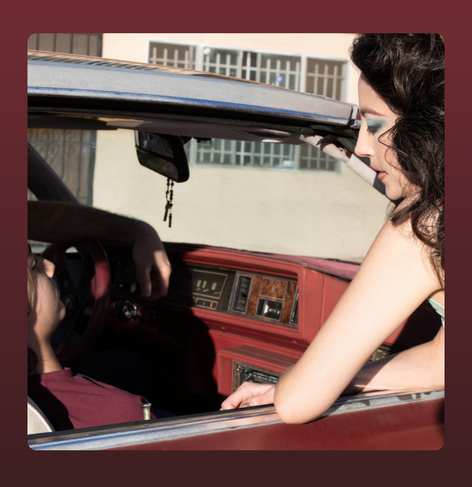 This image features a retro-styled scene with a woman leaning into a vintage red car, engaging with a man who is lounging in the driver's seat. The warm lighting and soft shadows create an intimate yet detached atmosphere, evoking themes of nostalgia, longing, or fleeting connection. The woman's dramatic makeup and the classic car interior suggest a cinematic quality, reminiscent of 1980s aesthetics.  

For Deafheaven's *Lonely People with Power*, this image could symbolize themes of isolation within relationships, the contrast of control and vulnerability, or the interplay of nostalgia and modern disillusionment. The framing emphasizes distance despite physical closeness, reinforcing the album's exploration of solitude and power dynamics.