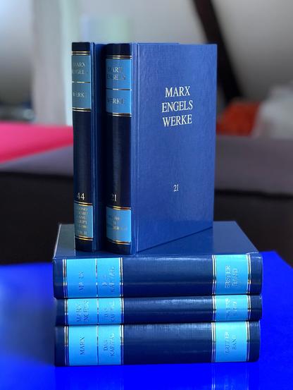 Aufeinandergestapelte Bände unserer Marx-Engels-Werke (MEW) in typischen Blautönen mit Gold-Prägung. Auf »Das Kapital« Band 1/2/3 stehen MEW Band 21 und 44.