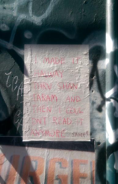 A sheet of white A4 paper is glued to an exterior wall that is covered in graffiiti. Handwritten on the sheet in all caps, in what looks like red crayon, is the following text: "I made it halway thru Shantaram and then I couldn't read it anymore." The words "Shantaram" and "couldn't" break onto the following lines, without hyphens. Below the red lettered text, in different handwriting, somebody has written "SAME!" in brown, also in all caps.