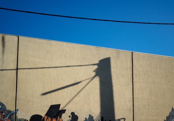 The shadow of a telephone pole cast on an exterior wall, and the powerlines and street light attached to the pole, intersect with lettering from a partially visible piece of street art in such a way that the shadow looks like the beak of a large bird that is feeding a smaller bird.