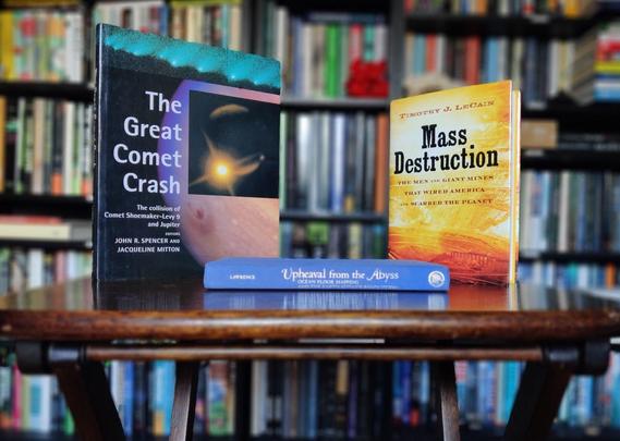 A photo of three books standing on a small, brown, wooden table. On the left, standing up, The Great Comet Crash, showing a picture of Jupiter taken with a telescope. In the middle, lying down, Upheaval from the Abyss, showing a marine blue spine with silver lettering (the dust jacket is missing). On the right, standing up, Mass Destruction, showing an golden/honey coloured aerial photo of an open-pit mine. The out-of-focus background shows black shelves full of books and part of the beech-coloured laminate floor.
