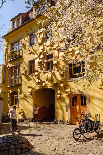 Das Bild zeigt eine außergewöhnliche gelbe Hausfassade mit wellenartig abstehenden Elementen, die den Eindruck erwecken, als würde die Fassade aus aufgerolltem Papier bestehen. Die Fenster sind von schlichten, dunklen Holzrahmen umgeben, während ein blühender Baum im Vordergrund die Szene mit zarten weißen Blüten schmückt.

Im Hof vor dem Gebäude sind zwei Personen zu sehen: Eine Frau mit heller Jacke und gemustertem Rock geht in Richtung des Hauses, während ein Mann mit Rucksack in der Durchfahrt telefoniert. Rechts steht ein blaues Lastenrad mit Kindertransport-Aufsatz. Das Sonnenlicht und die Schatten der Äste verleihen der Szene eine warme und lebendige Atmosphäre.