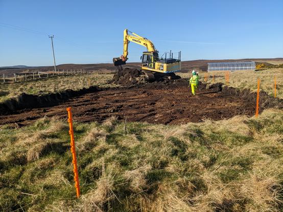 A photo of an open field with a large yellow digger excavating the ground. There's a large area where the top soil has been removed. A person in high visibility clothing is observing the activity and orange poles sticking in the ground mark out the space.