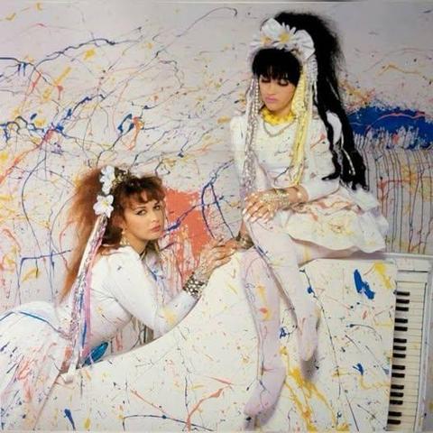 A white baby grand piano lies on its side, Rose McDowall casually sits on top, Jill Bryson leans on to it. Both people are dressed in white and they, the piano and the wall are splattered with coloured paint