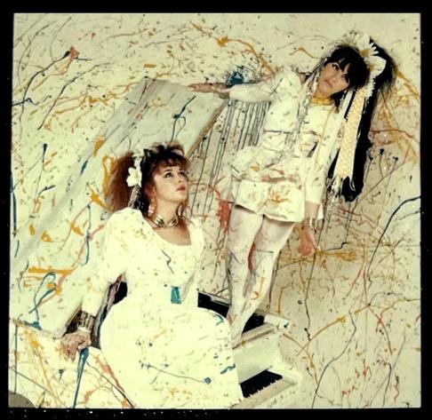 Rose stands and Jill sitson top of a white baby grand piano, dressed in white. The whole scene is splattered with coloured paint.