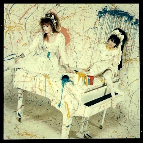 Rose and Jill sit on top of a white baby grand piano, dressed in white. The whole scene is splattered with coloured paint, and open paint tins are on top of the piano