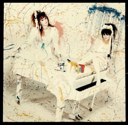 Rose and Jill sit on top of a white baby grand piano, dressed in white. The whole scene is splattered with coloured paint, and open paint tins are on top of the piano