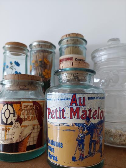 Auf dem Bild sind sechs Aufbewahrungsgläser zu sehen, drei Spaghettigläser mit Korkdeckel, im Vordergrund eines mit einem alten Aufdruck "Au petit Matelot" und eines mit "Perles d"Huile de Bain".